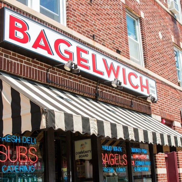 An outside view of the storefront Bagelwich Bagel Bakery & Deli - 652 Bloomfield Ave. There is a large sign in red lettering that reads BAGELWICH and a black and white striped awning underneath that sign. The entrance is partially visible. The building is red brick, and the weather is bright and sunny.