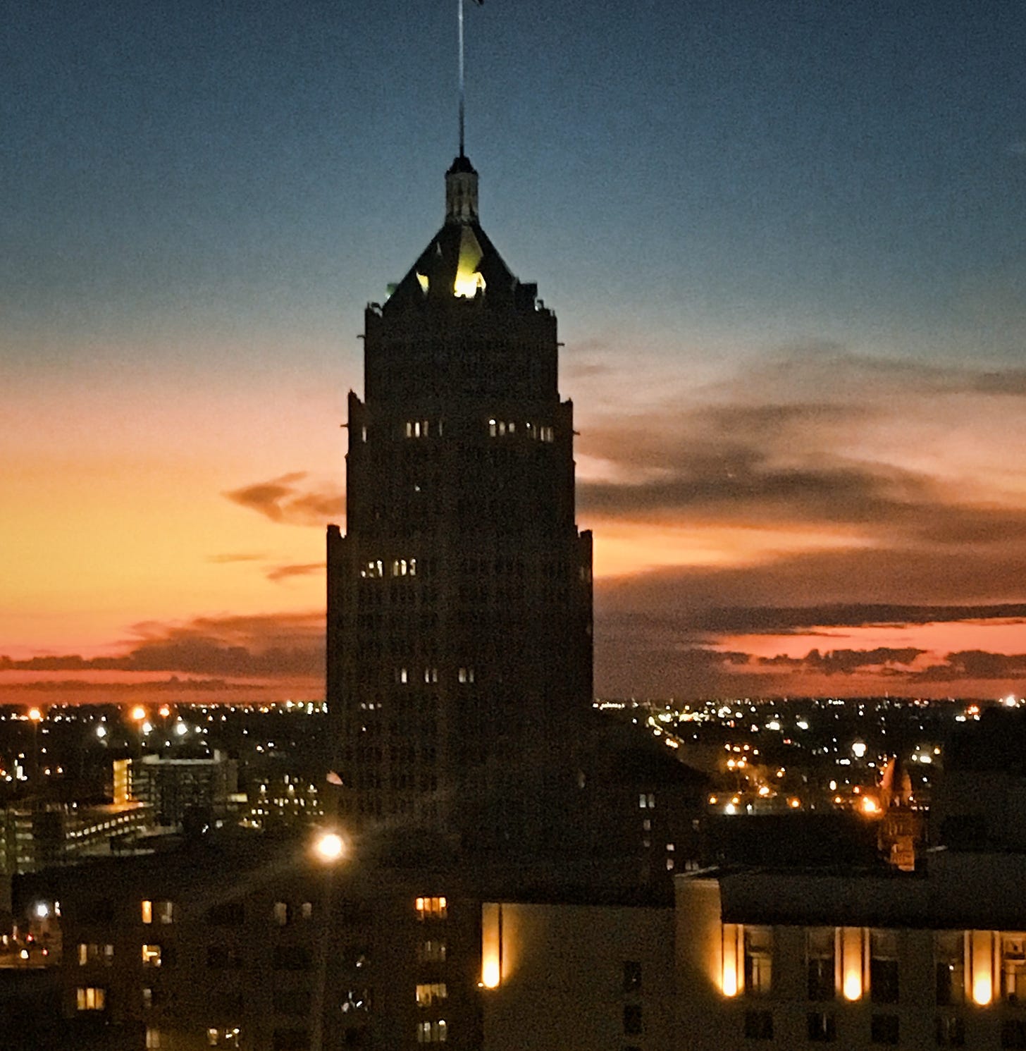 Orange and yellow streaks in the sunset with dark blue-grey sky with the city lights and a single tall building in the middle of the scene