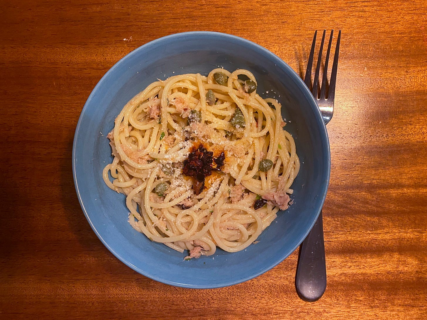 A blue bowl of spaghetti with tuna and capers, sprinkled with parmesan and with a blob of chili crisp in the centre. A fork is on the table to the right of the bowl.