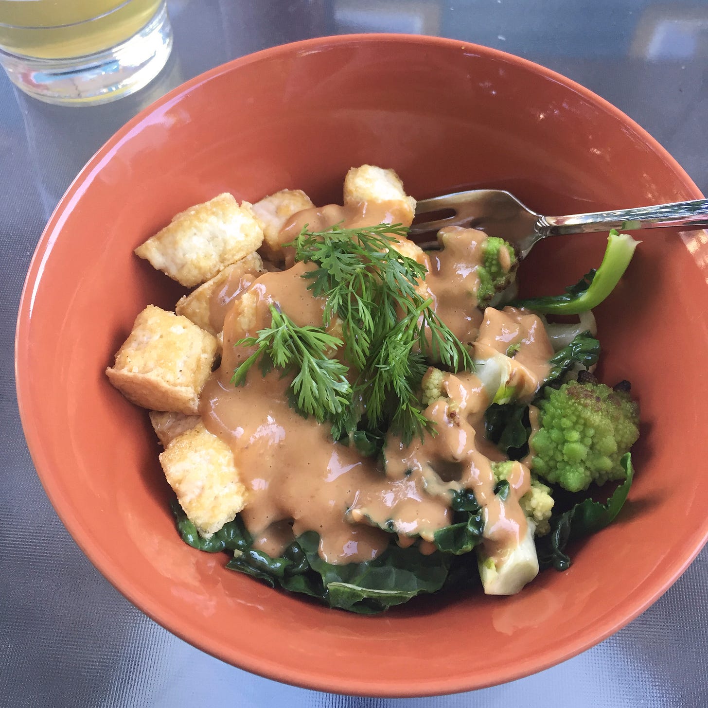 An orange bowl with romanesco broccoli, kale leaves, and crispy pieces of tofu with peanut sauce and cilantro overtop.