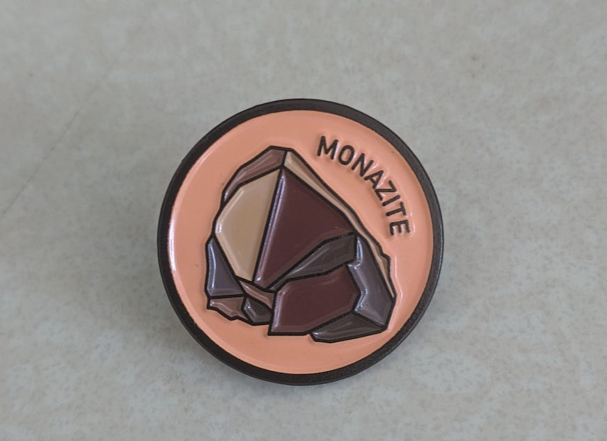 An enamel pin with an illustration of a reddish-brown rock against a salmon-ish background. The word "monazite" curves around the right side of the rock.