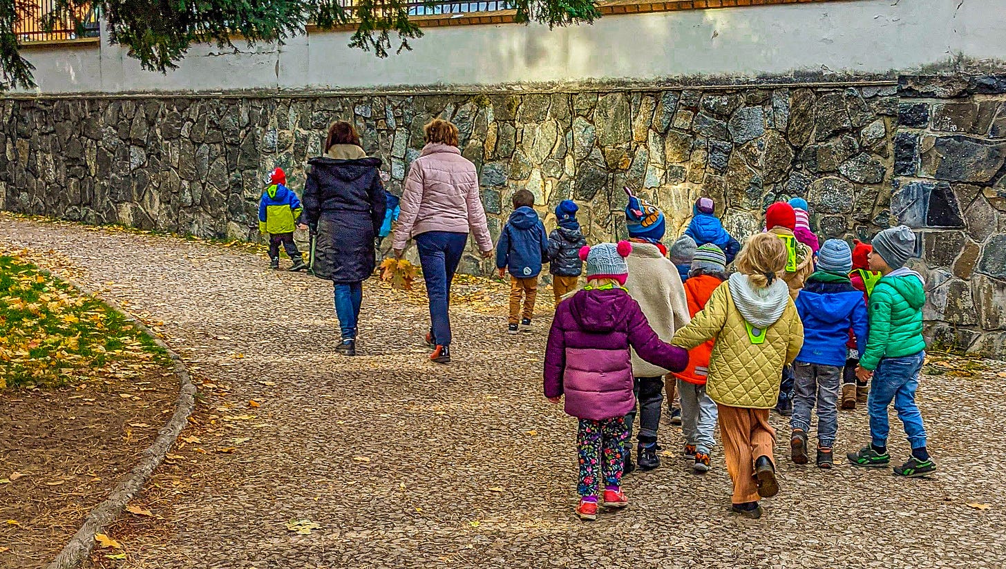 A group of very young children holding hands and walking through a park with their adult guardians. 