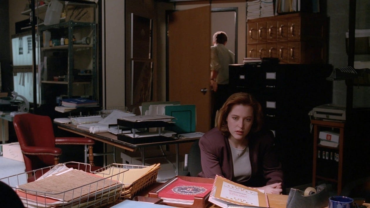 Scully looking annoyed in a messy office while Mulder walks out of the door behind her