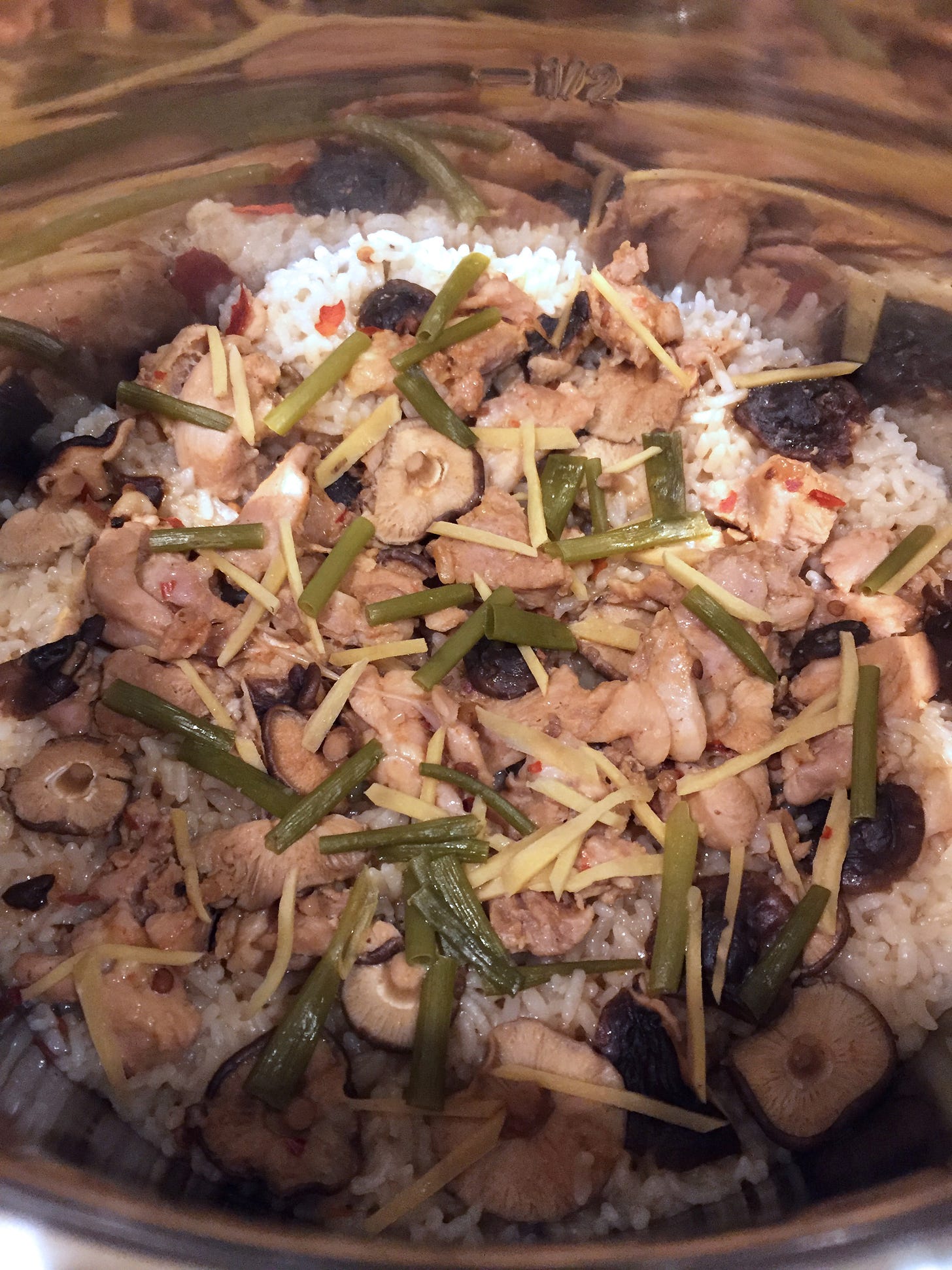 cooked chicken pieces and small shiitake mushrooms sit on a bed of rice in a large silver pot. Slices of ginger and green onion are scattered on top.