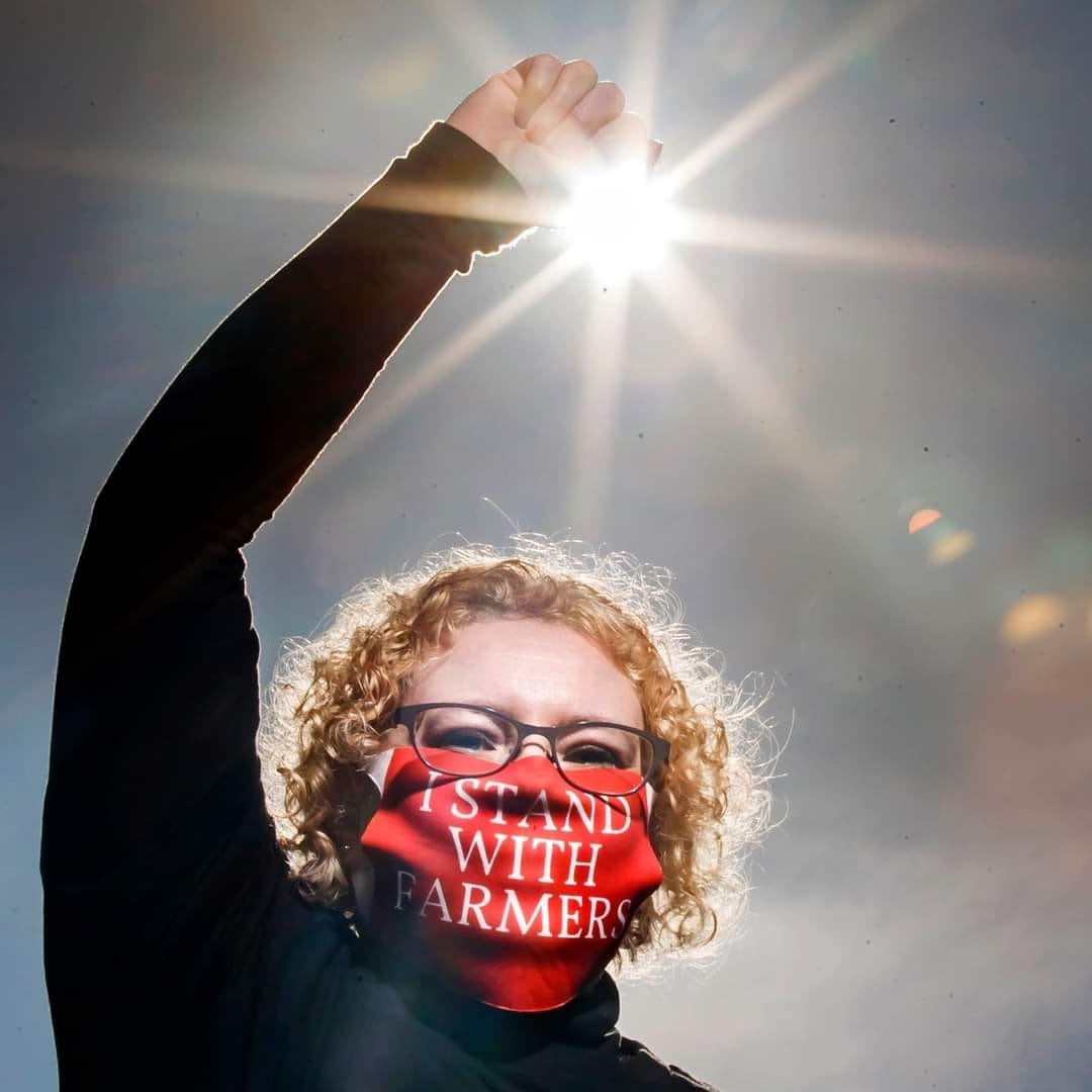 Joanna Pollard holds a fist up to the camera, silhouetted by the sun. She has a mouthmask on that reads 'I stand with farmers.' She is wearing a black jumper and glasses.