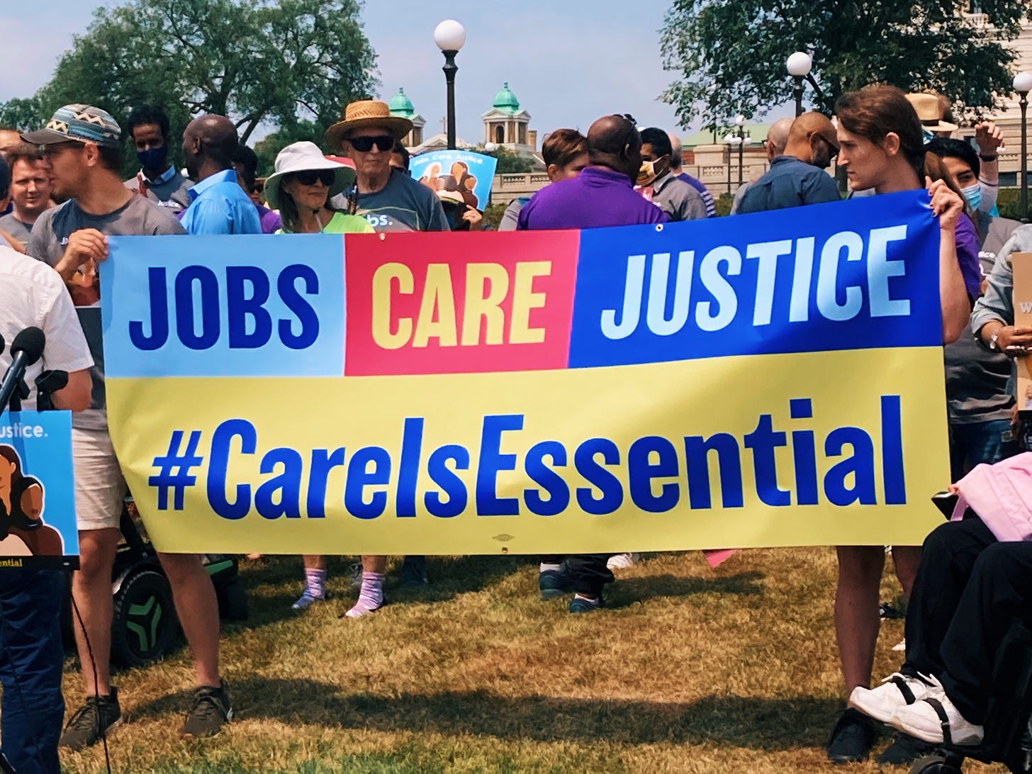 people hold up banner with text reading "jobs care justice #careisessential" in blue and yellow text with yellow, red, and blue background