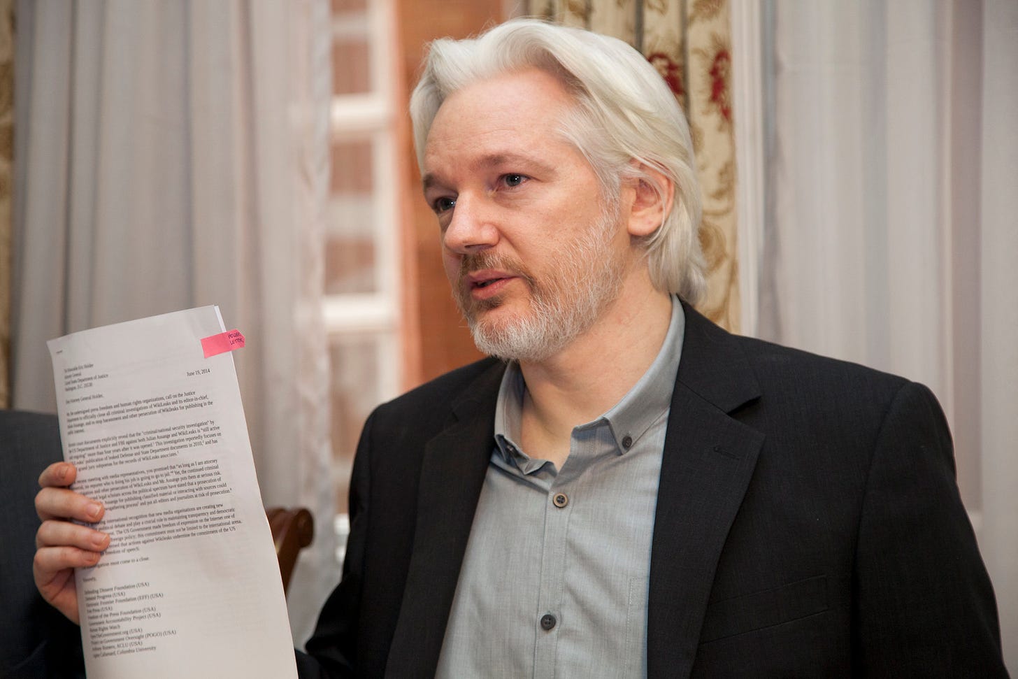 WikiLeaks co-founder and publisher Julian Assange holds a stack of documents during a press event. He's wearing a gray shirt and black suit coat.