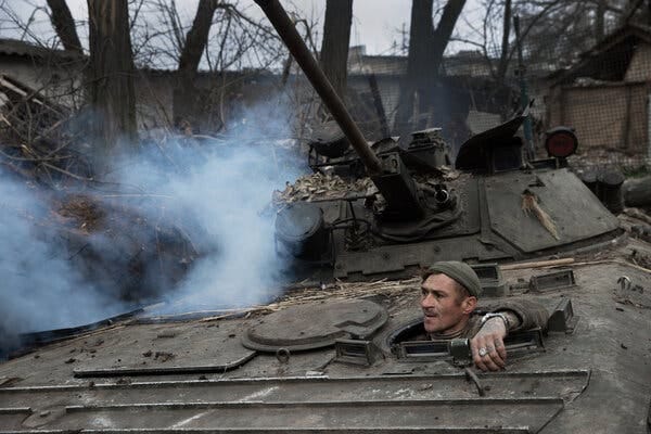 A Ukrainian soldier peeking out of a tank with smoke and trees behind him.