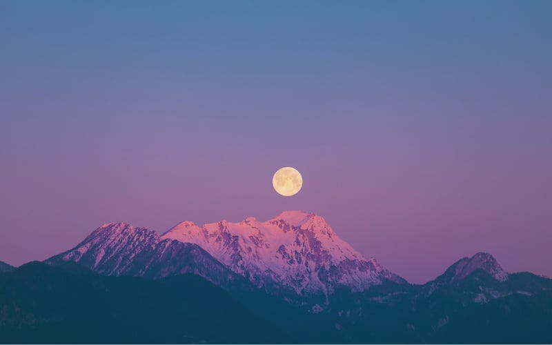 A full moon rises above a snow-capped mountain peak. The sky is a dusky blue and violet, it's not yet nighttime.