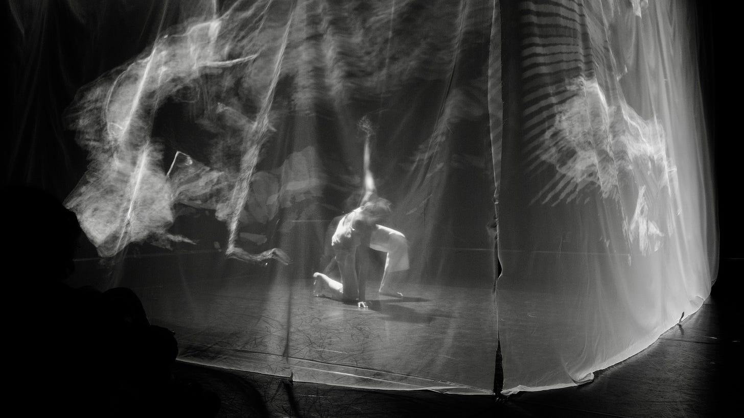 A long exposure black and white photo of Yo-Yo Lin in a blur, dancing inside a translucent fabric cube. White projections appear like apparitions on the fabric walls. She is on one knee, her head down, arm up in the air holding onto black wires hanging from the ceiling. 