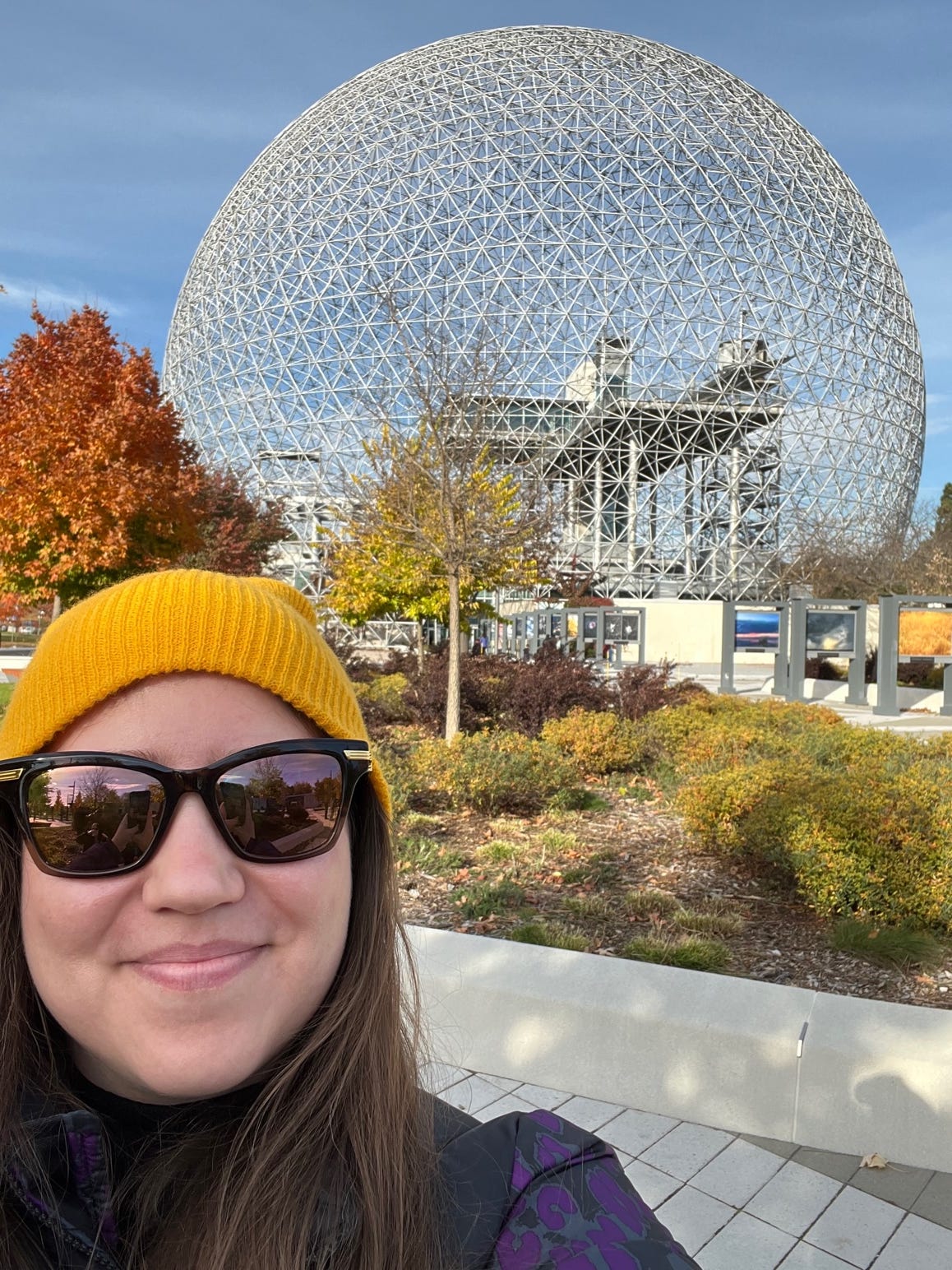 A selfie of a girl with brown hair, a yellow hat, and a purple jacket wearing sunglasses in front of the Biosphere of Montreal