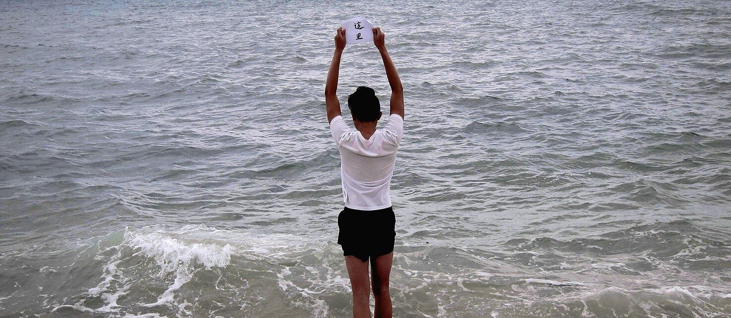 jee chan standing in the waves at East Coast Park, holding up a piece of calligraphy written by their grandmother/婆婆 that reads 這里, meaning “here”