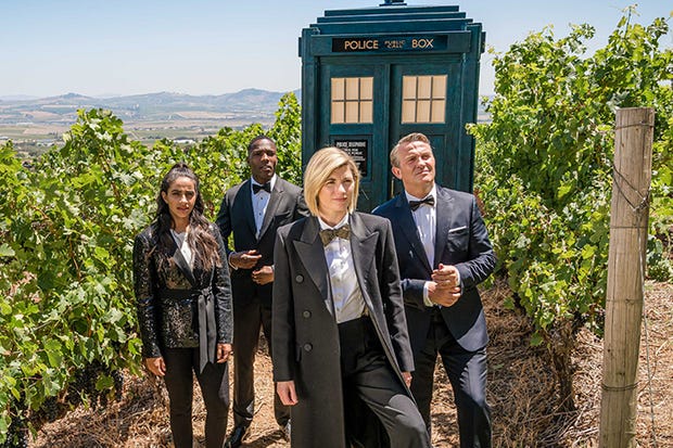 This image shows Team TARDIS dressed in tuxedos while standing in front of the TARDIS before entering the estate of Daniel Barton. From left to right stand Yasmin "Yaz" Khan (played by Mandip Gill), Ryan Sinclair (played by Tosin Cole), the Thirteenth Doctor (played by Jodie Whittaker), & Graham O'Brien (played by Bradley Walsh). All four plus the TARDIS stand in a vineyard and look off camera at Barton's unseen villa. This image shows Team TARDIS dressed in tuxedos while standing in front of the TARDIS before entering the estate of Daniel Barton. From left to right stand Yasmin "Yaz" Khan (played by Mandip Gill), Ryan Sinclair (played by Tosin Cole), the Thirteenth Doctor (played by Jodie Whittaker), & Graham O'Brien (played by Bradley Walsh). All four plus the TARDIS stand in a vineyard and look off camera at Barton's unseen villa.