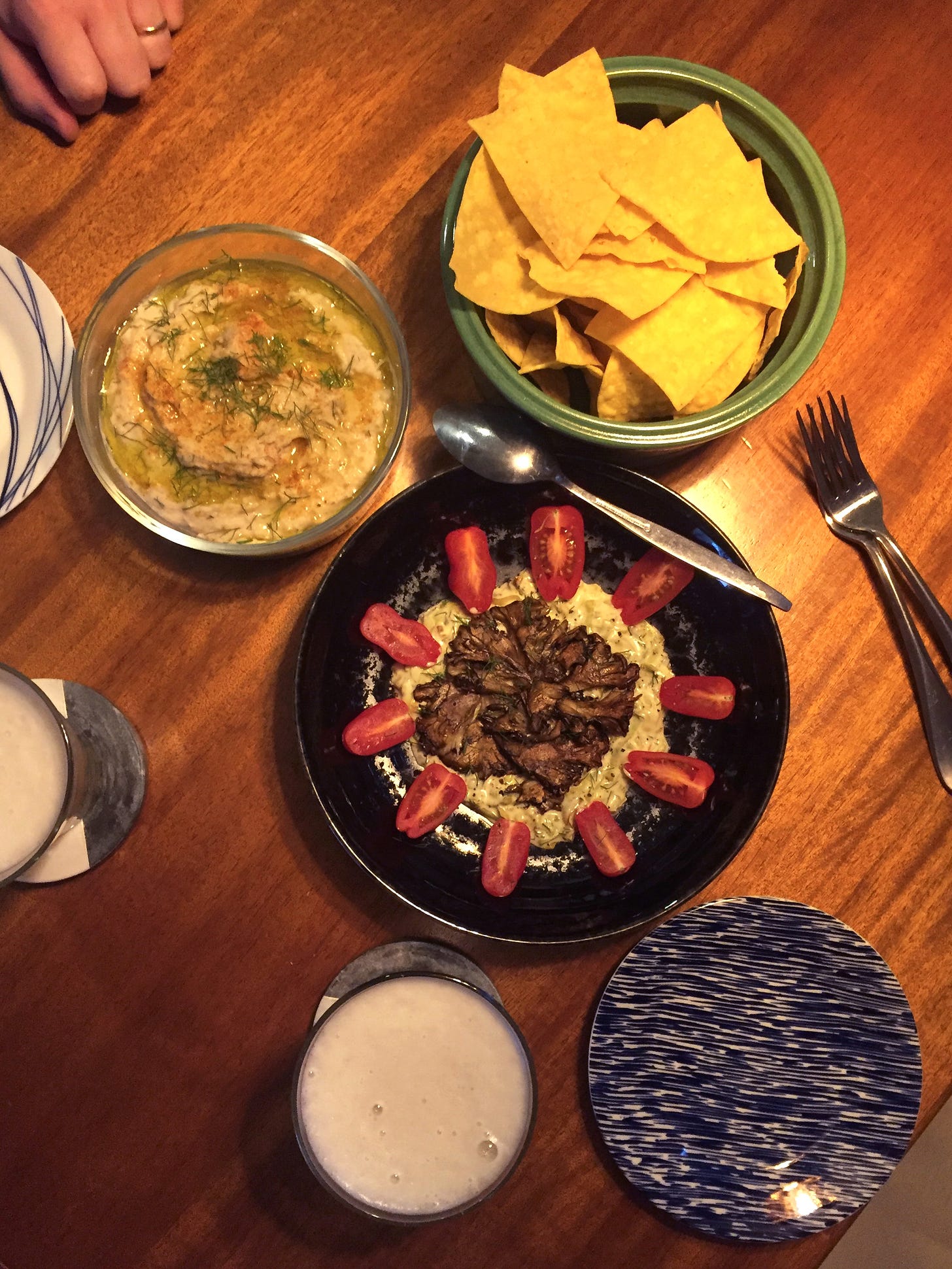 From above, a table with an arrangement of dishes: a glass bowl of creamy bean white bean dip, a green ceramic bowl of tortilla chips, and below them, a black shallow dish with maitakes resting on top of the remoulade, with small tomatoes around the edge.