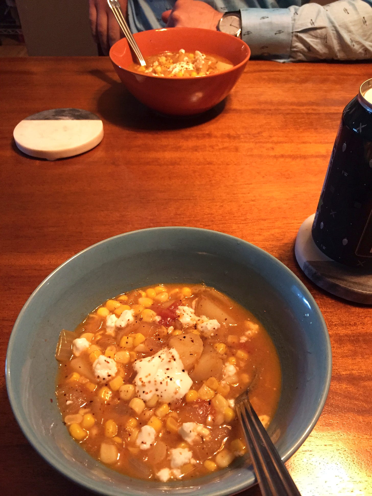 Two bowls of corn chowder across from each other on the table. On top of the soup is a dollop of yogurt, crumbles of feta, and tajín seasoning. On a coaster next to one of the bowls is a blue can of beer.