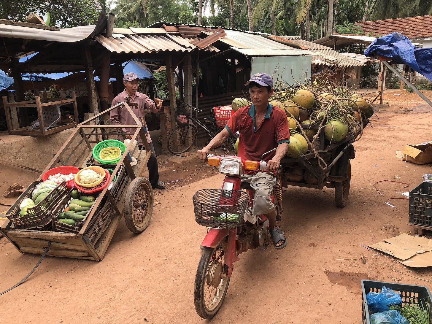 Two men with food delivery. One with a hand cart full of fresh vegetables; the other on a scooter with a trailer full of what looks like melons. No packaged food in sight.