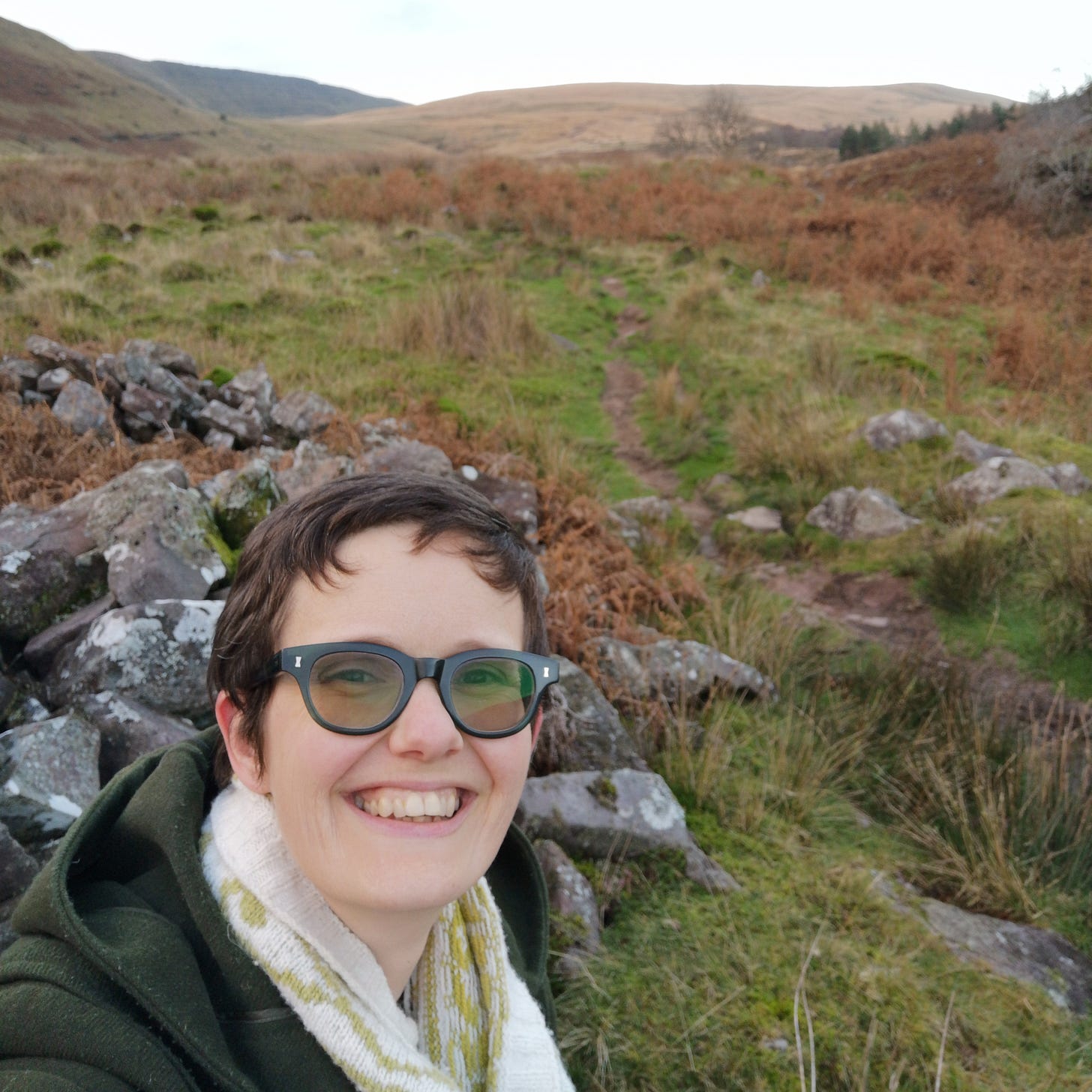 Image description: A selfie of Katie grinning at the camera in front of a scrubby walk up the valley.