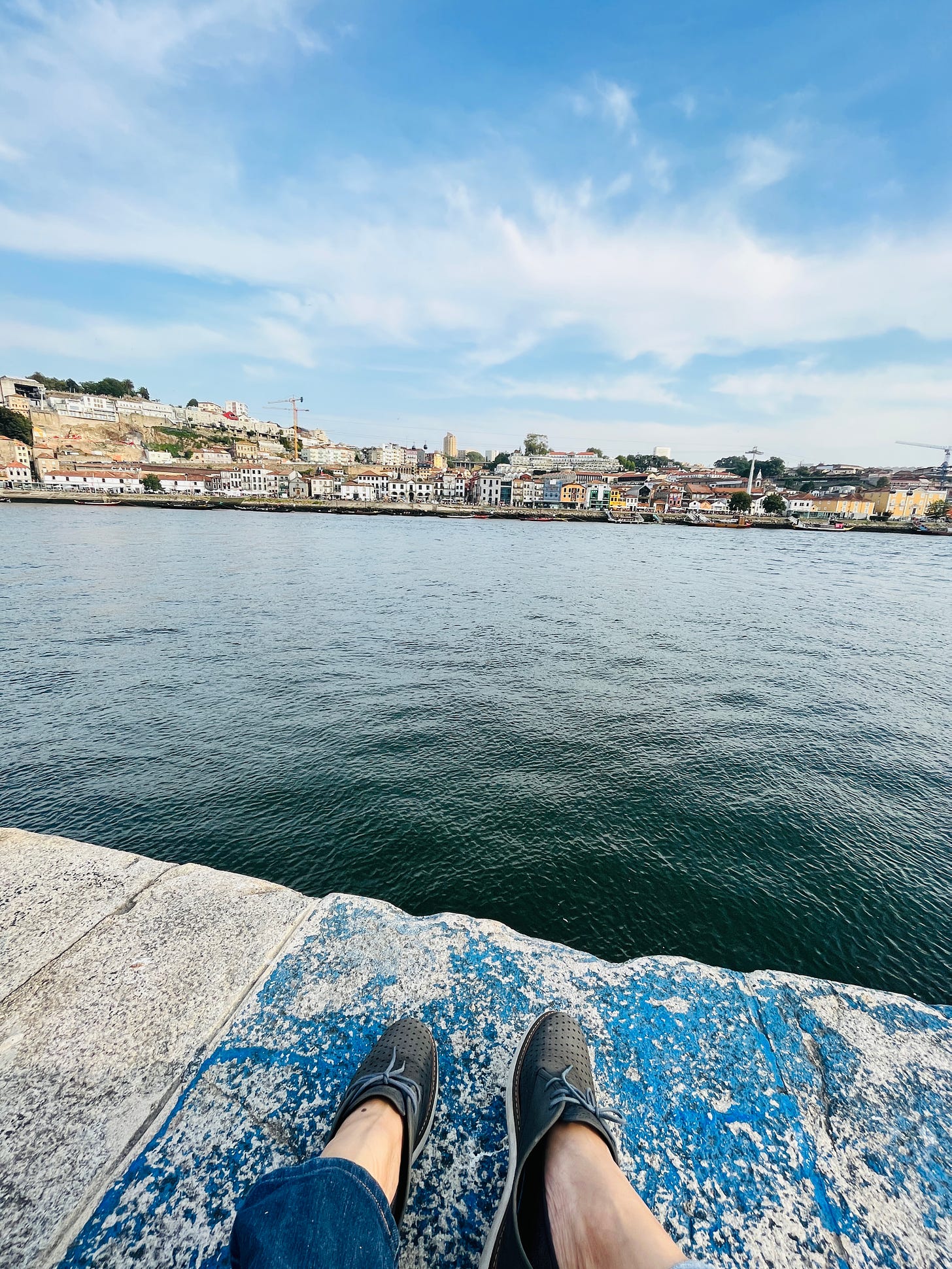 Image: photo of the Duoro river with the Vila Nova de Gaia opposite. In the foreground, there are the writer’s stretched-out feet clad in her trusty Clarks, relaxing in the moment.