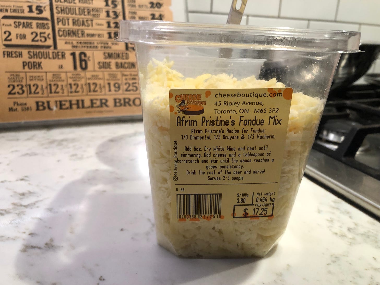 A container of shredded cheese from Cheese Boutique on a kitchen counter. The label reads, "Afrim Pristine's Fondue Mix - Afrim Pristine's Recipe for Fondue: 1/3 Emmenthal, 1/3 Gruyere & 1/3 Vacherin. Add 6oz. Dry White Wine and heat until simmering. Add cheese and a tablespoon of cornstarch and stir until the sauce reaches a gooey consinstency. Drink the rest of the beer (sic) and serve! Serves 2-3 people. 