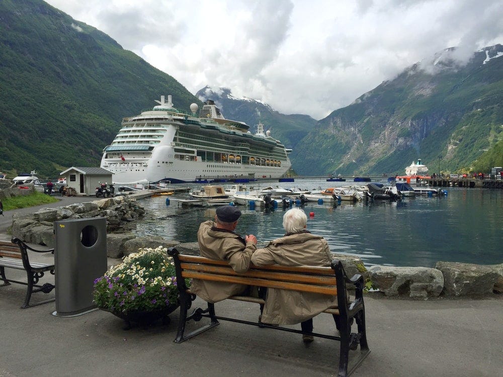 woman in brown coat sitting on brown wooden bench near white cruise ship during daytime, Julius Yls