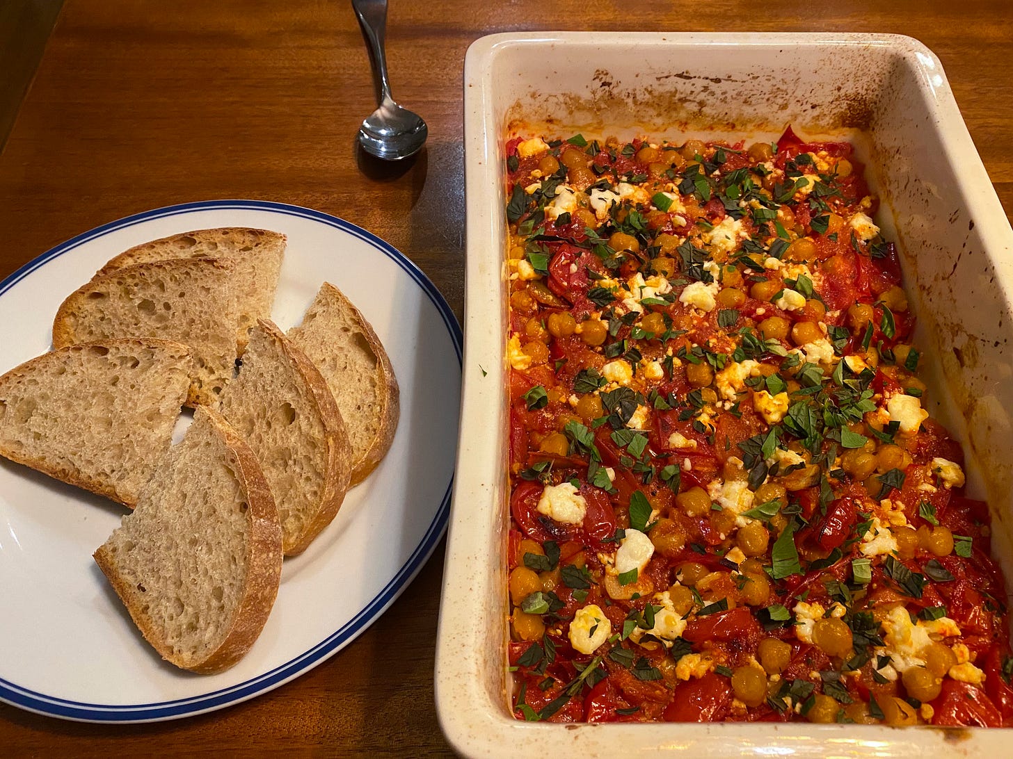 In a white rectangular baking dish, a mixture of baked chickpeas and diced tomatoes, covered with large crumbles of feta and chopped fresh herbs. To the left of it is a plate with three slices of sourdough cut in half.
