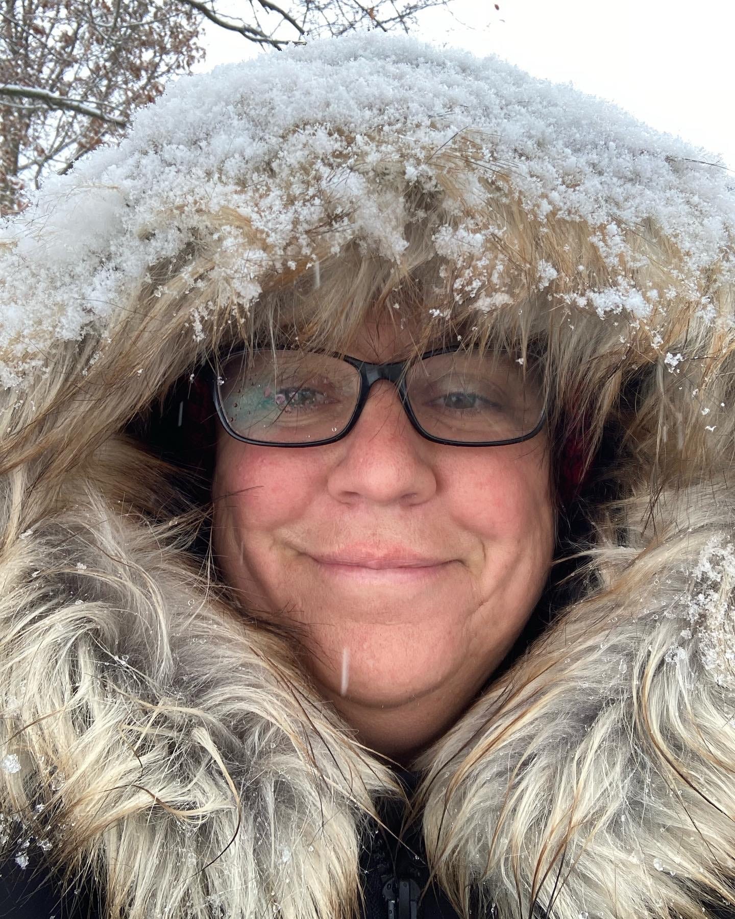 Robyn in a winter coat with the fur-lined hood pulled up and covered in snow and smiling, but not with teeth, never with teeth