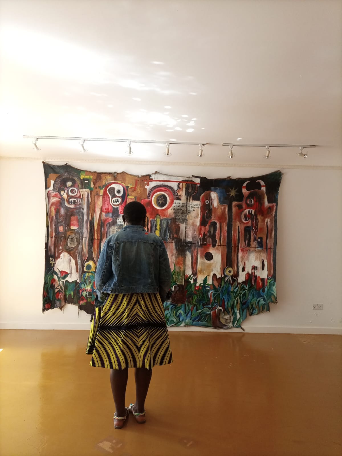 Black person with short hair wearing yellow and purple dress, denim jacket and sandals standing in front of a large painting