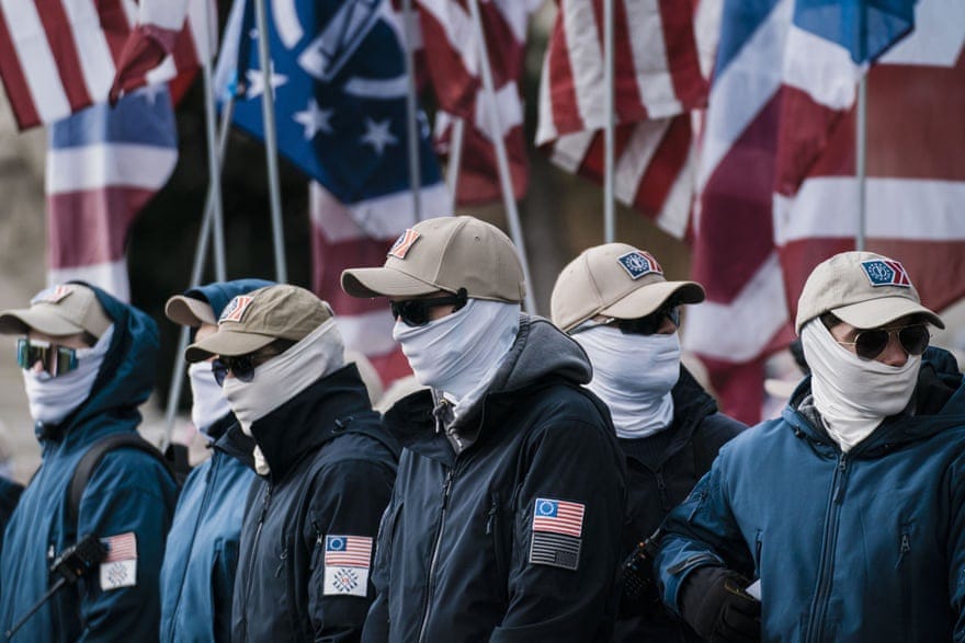 Group of men wearing balaclavas covering their faces and khaki colored caps stand in front of a row of US flags