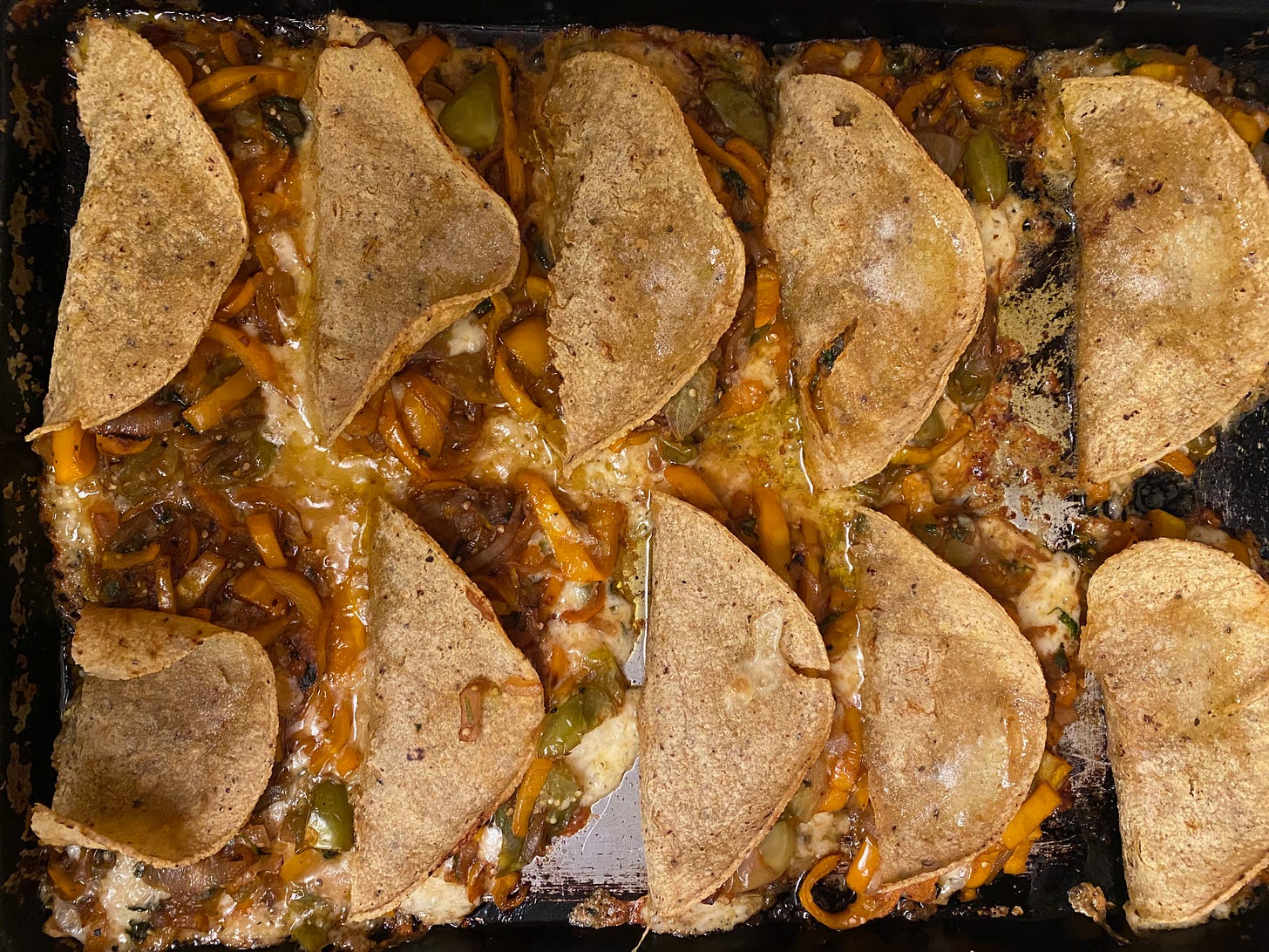 A tray of ten baked quesadillas. Half-moon shaped corn tortillas slightly overlap on the pan, the filling is oozing out all around them: slices of yellow peppers, pools of butter, and partially melted cheese.