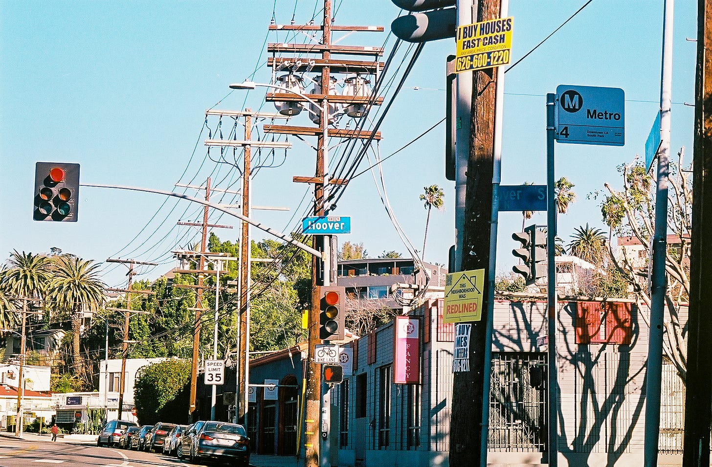 Photograph of the intersection of Santa Monica Blvd and Hoover with businesses, trees, and traffic signals. On the right hand side of the frame is a wooden pole with a sign that says “I Buy Houses Fast Cash” on the top and one of my signs on the bottom. 