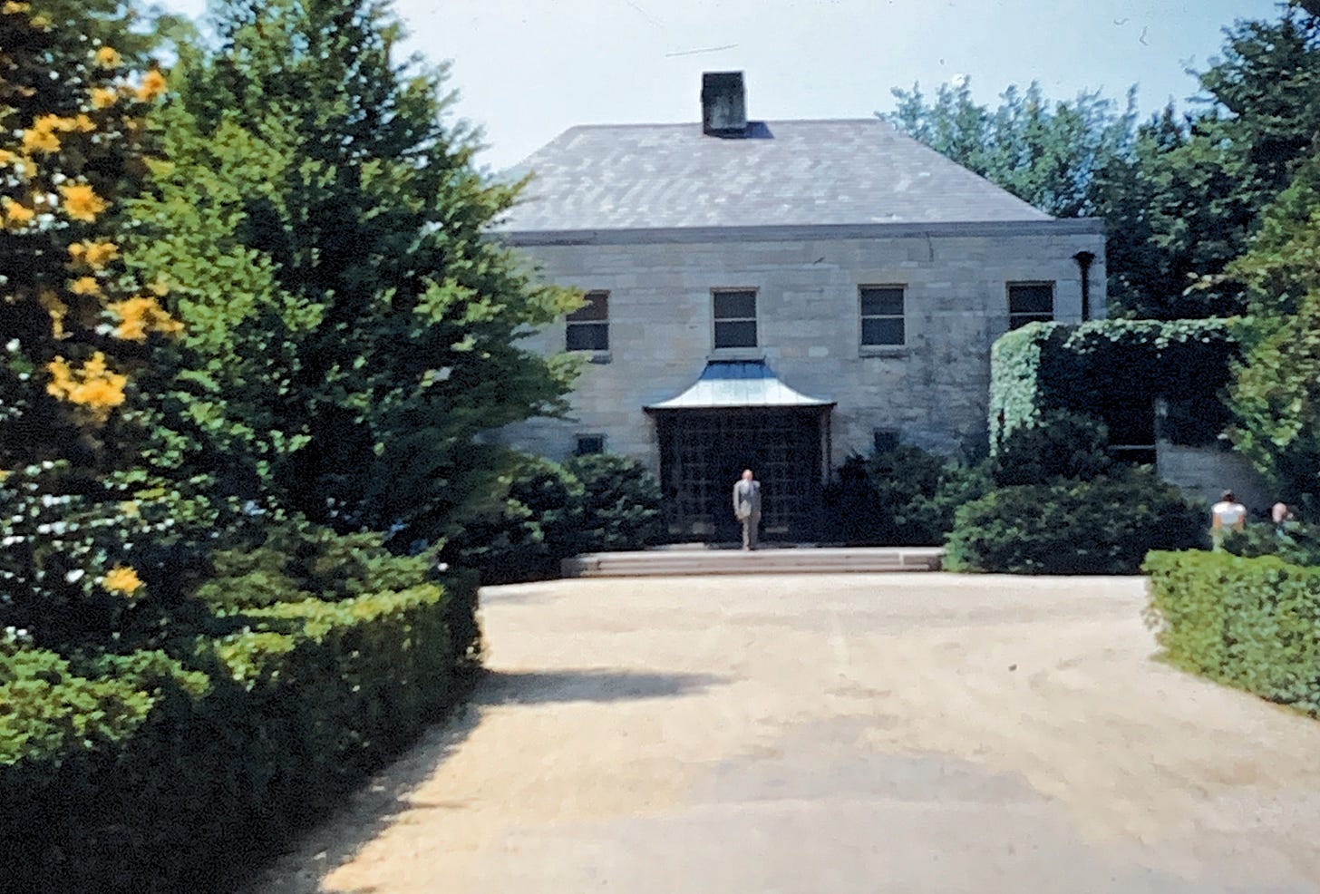 A white stone building with a small archway door, with hedges leading up to it and a man walking down the front steps.