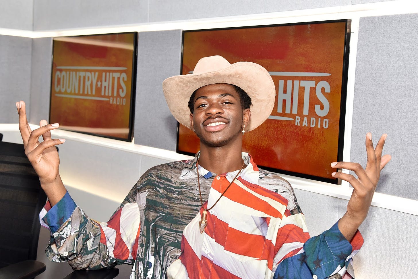 Photo of Lil Nas X wearing an American flag button down and cowboy hat, sitting in front of two televisions that display a "Country Hits Radio" emblem.