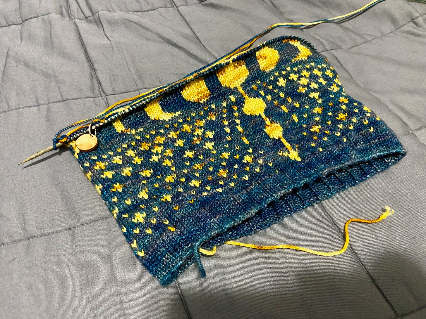 A partially knitted Ixchel Cowl, which features a blue backdrop and yellow stars, moons of varying sizes, and an arrow running down the center.