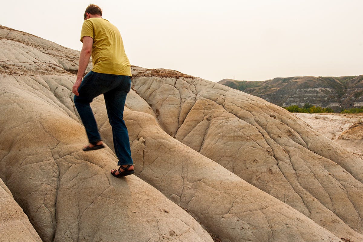 man in yellow shirt and chaco sandals walks up the slope of a small badlands crest, the sky murky with smoke