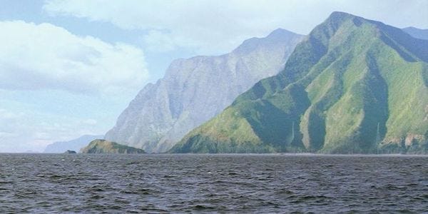 Picture of the island seen from the ocean. Its green mountains rise up to meet the clouds.