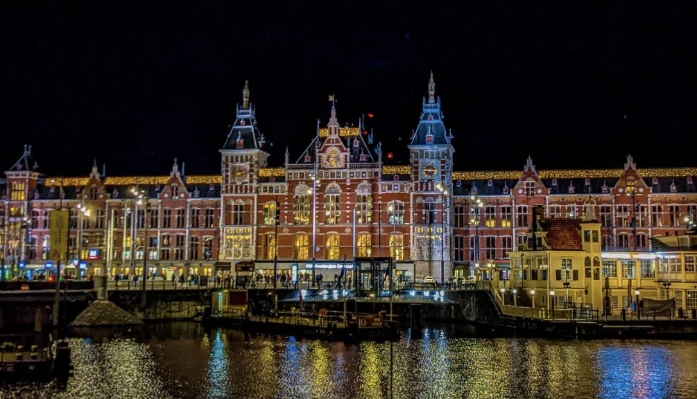 Amsterdam's central train station at night. A brick building with several towers and lots of windows, the yellow light reflected in the water. 