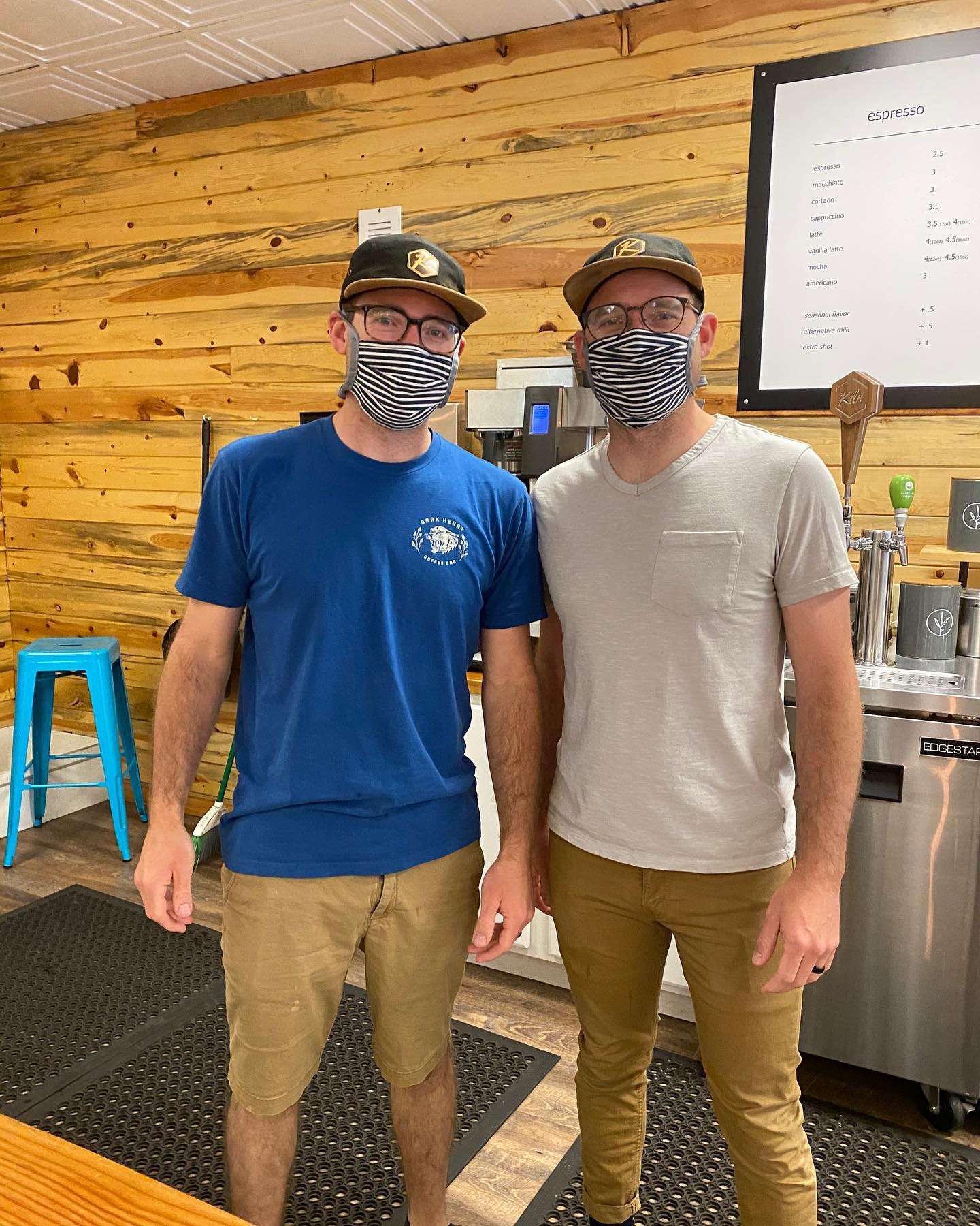 Identical twin brothers (caucasian) pose for the camera in matching masks and glasses in front of a pine wood wall at a coffee shop.