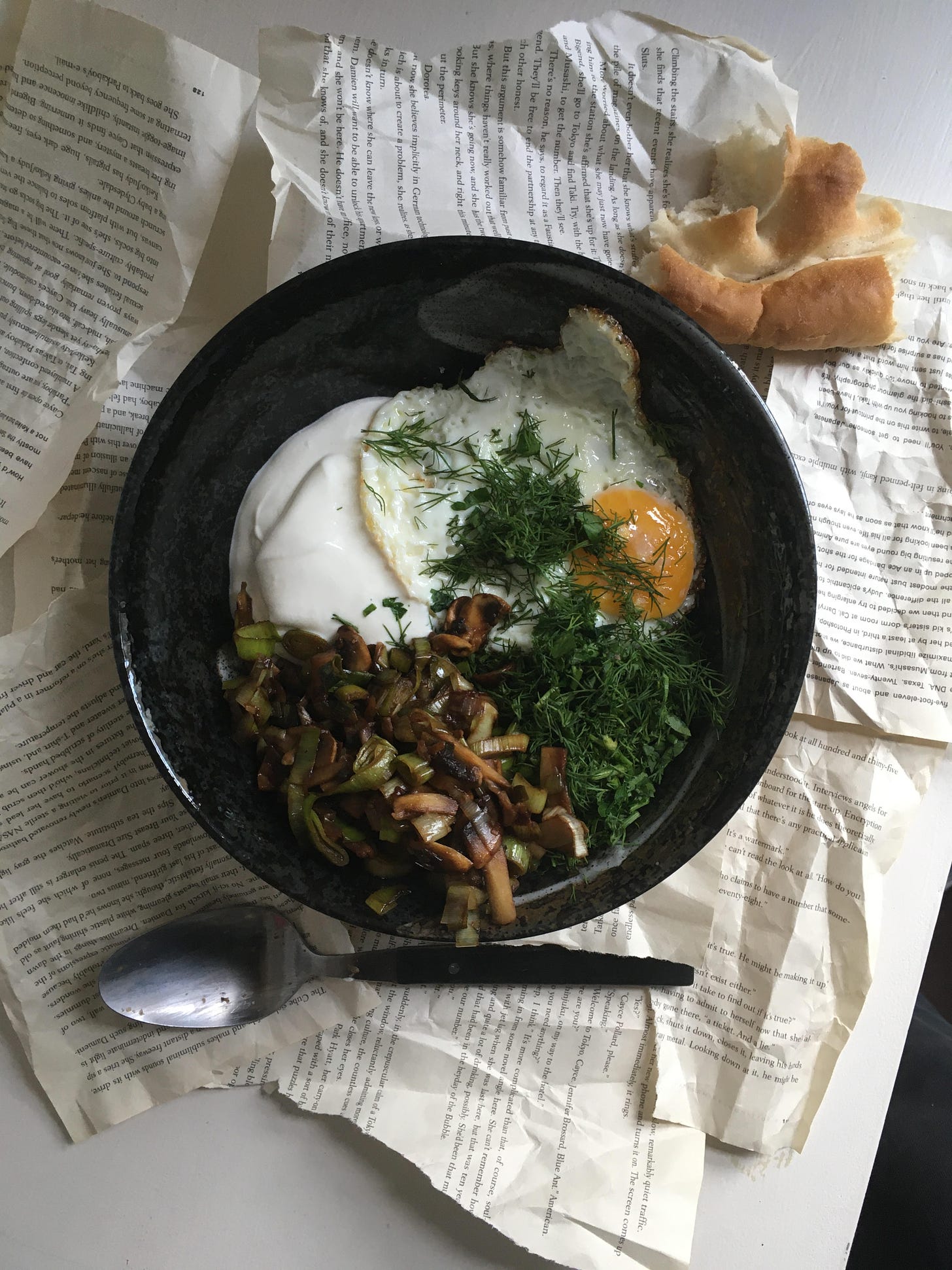 A black bowl with a fried egg, some herbs scattered over, chopped fried leeks and mushrooms, some sour cream. On the table around it, a spoon, a hunk of bread, and scattered pages torn from a book. 