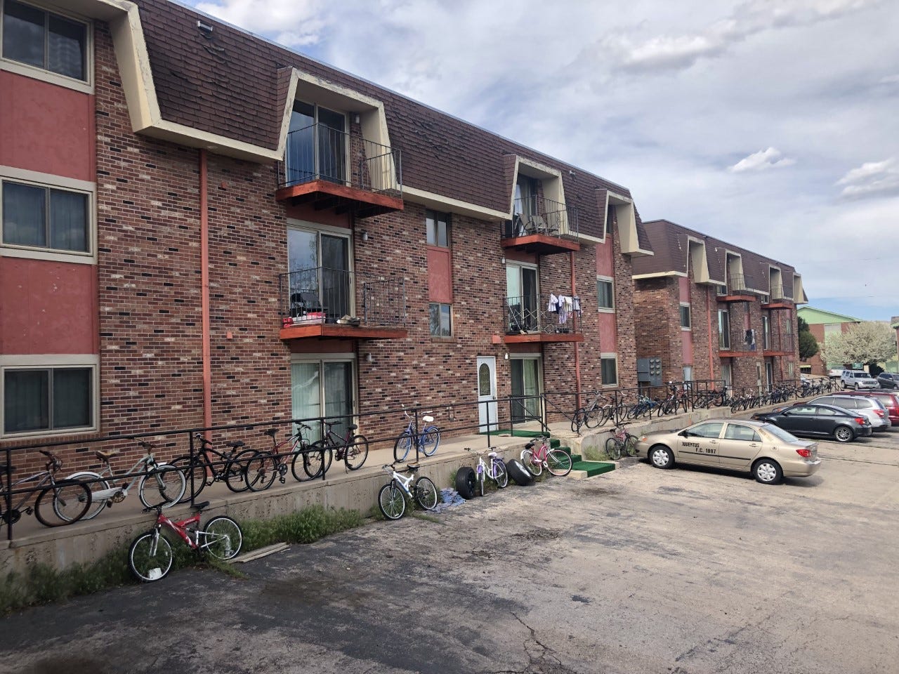 Two buildings, both brick and three stories tall, sit before a gray, cloudy sky. Bicycles and cars clutter the foreground.