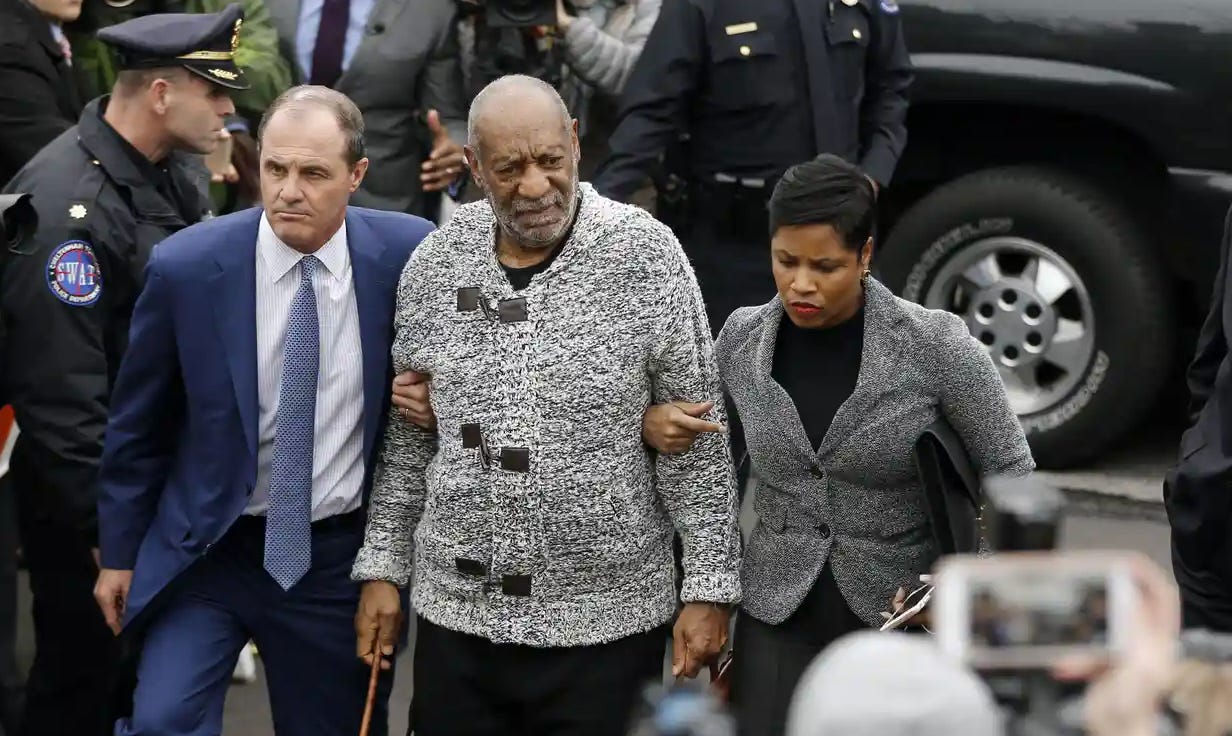 A picture outside of an elderly Black man standing centered wearing a grey sweater. He is aided on either side by younger people holding his arms. Behind are standing police officers in front of a car.