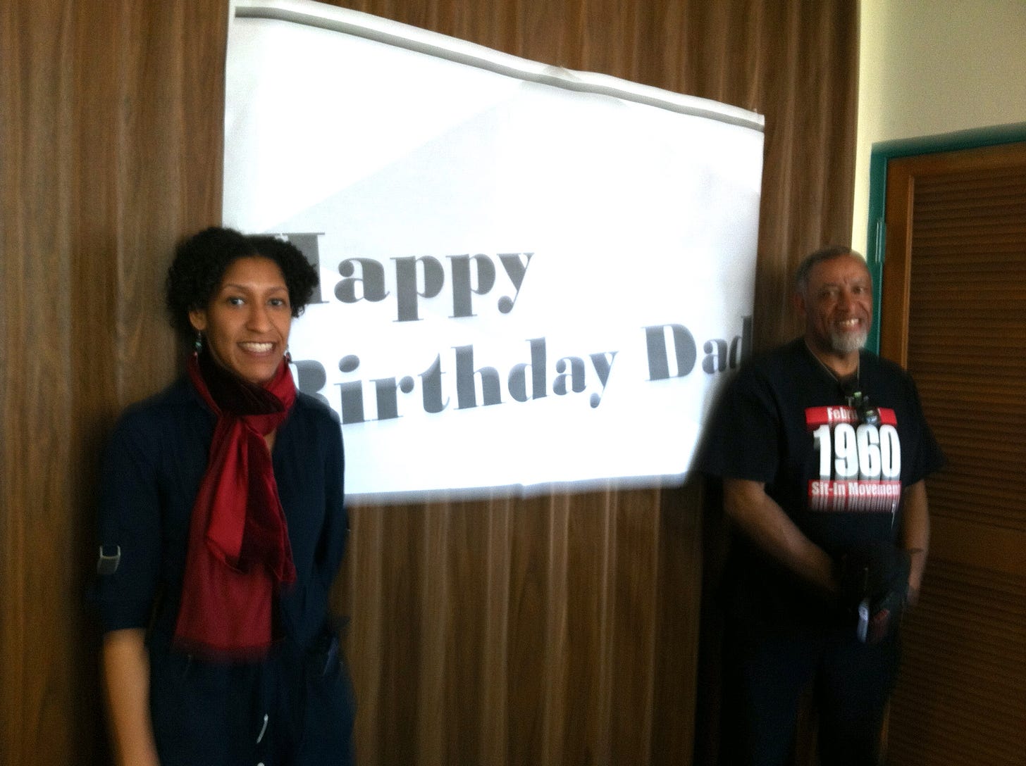 Kristen and her dad are standing on two sides of a white banner. The banner says Happy Birthday Dad!