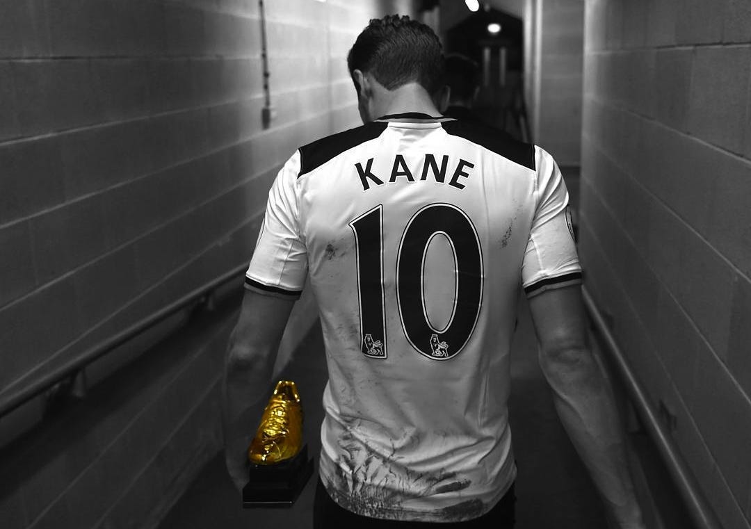 Black and white image of Harry Kane wearing a Tottenham jersey. He is walking down a hallway with the Golden Boot in his left hand.