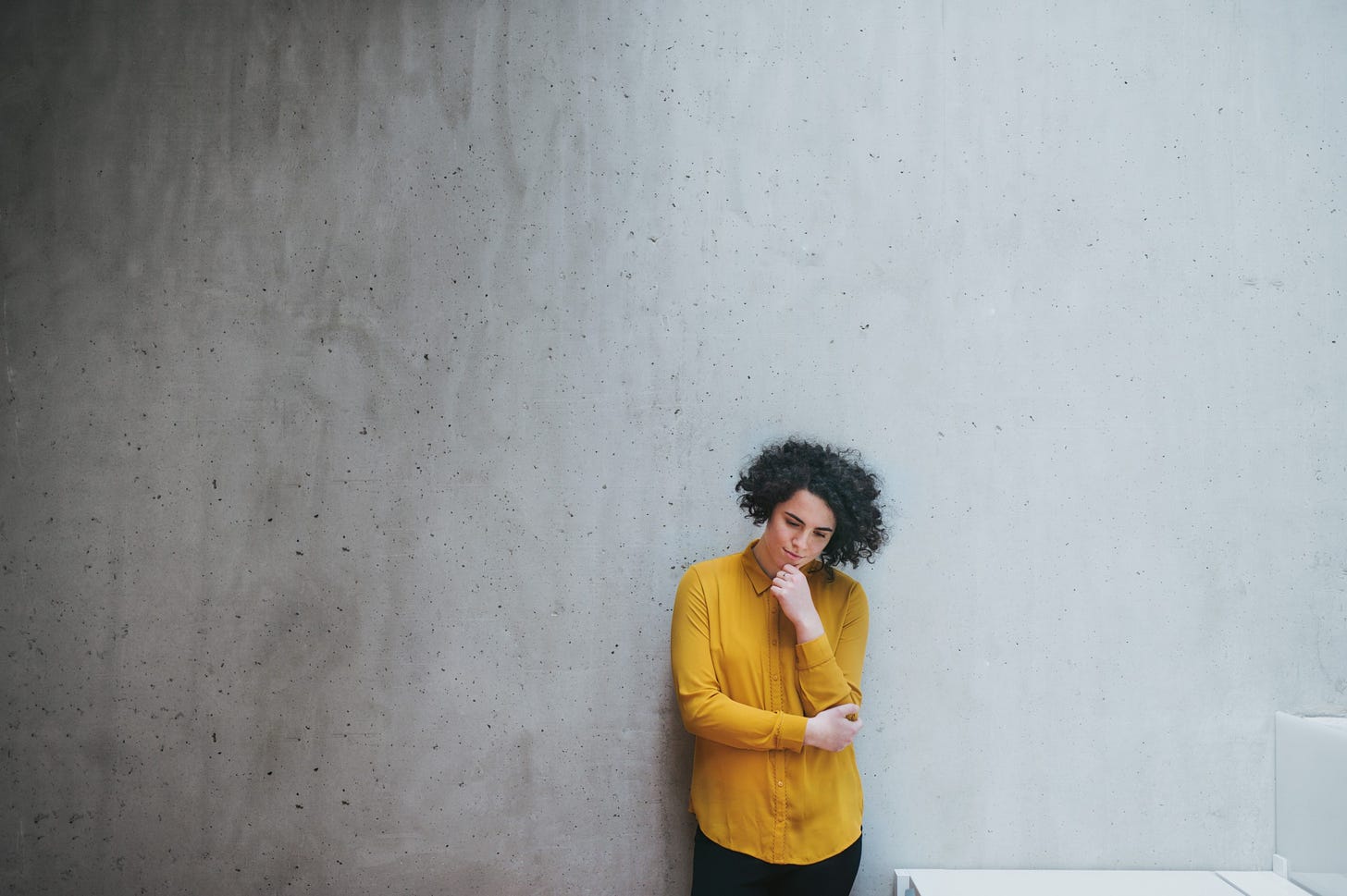 A woman wit curly hair and a yellow shirt stands against a bare concrete background pensively.