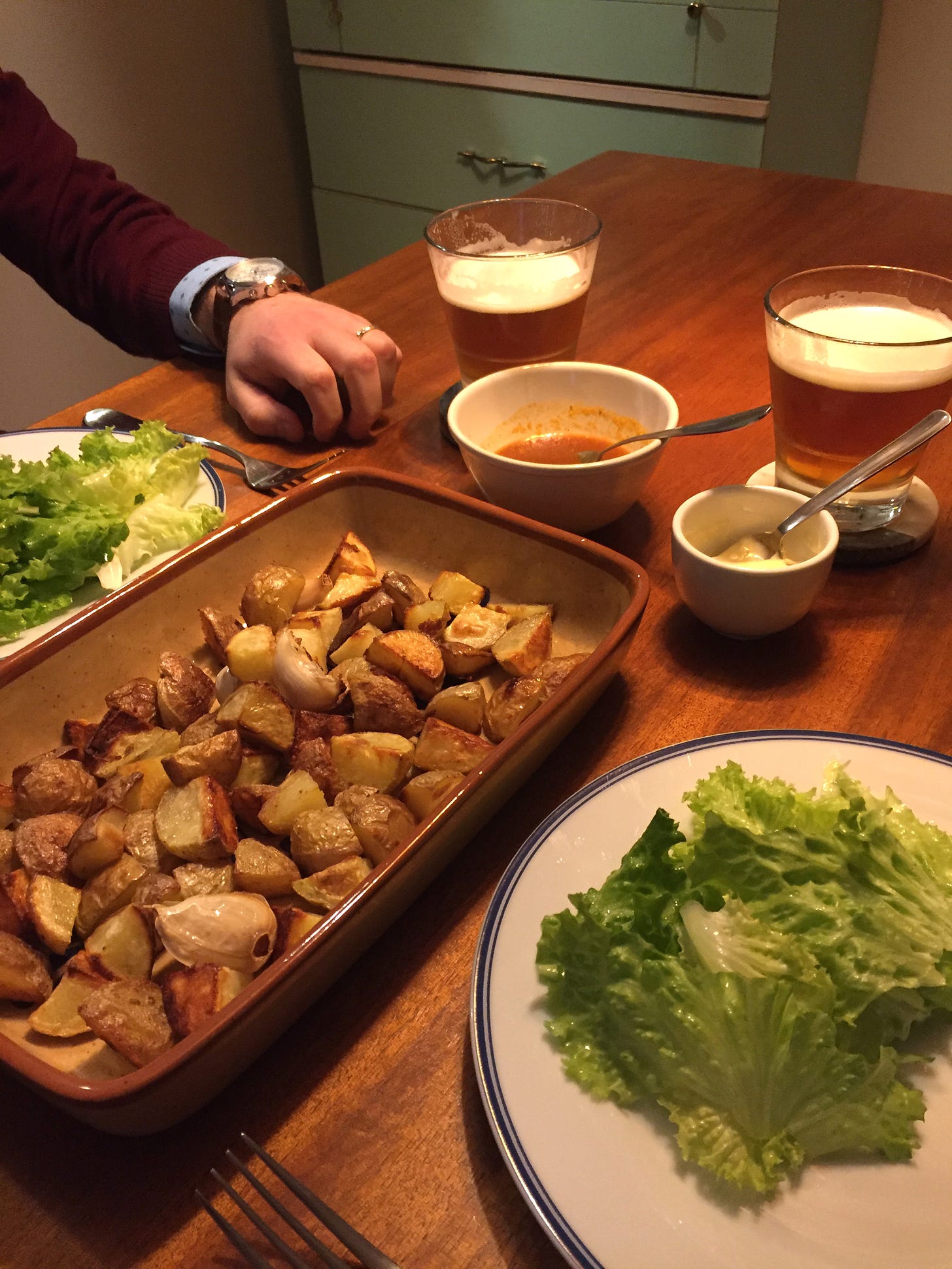 a square pan full of crispy potato pieces and whole cloves of roasted garlic. Two plates of green salad sit opposite. In the background are a dish of salsa brava and a dish of garlic mayo with spoons in them, and two glasses of beer on coasters.