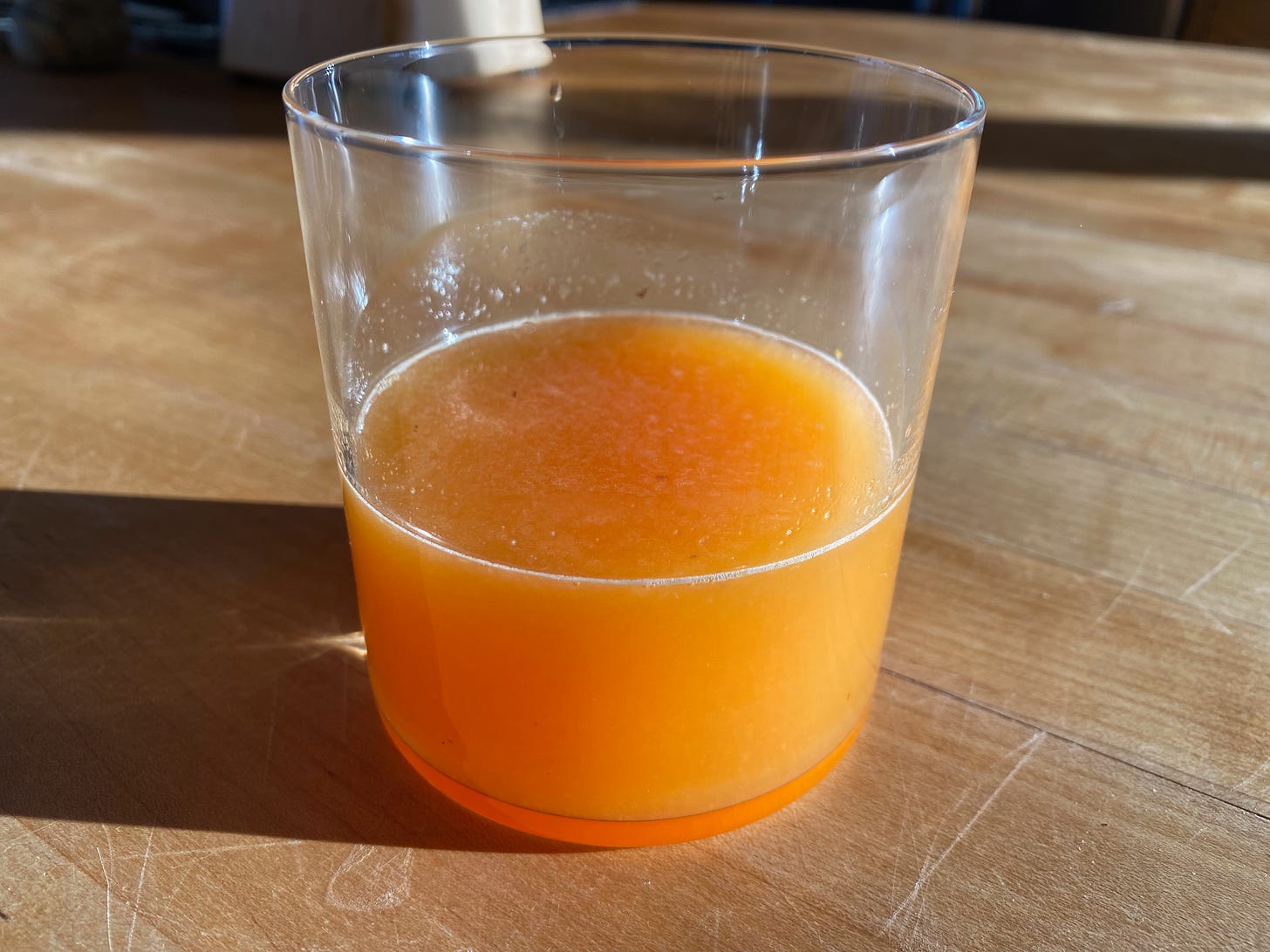 A class of freshly-squeezed orange juice sits on a sunny wooden countertop.