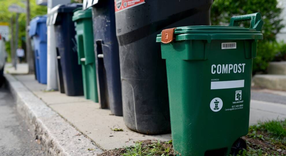 A group of black, green and blue trash and compost bins sit on a sidewalk.