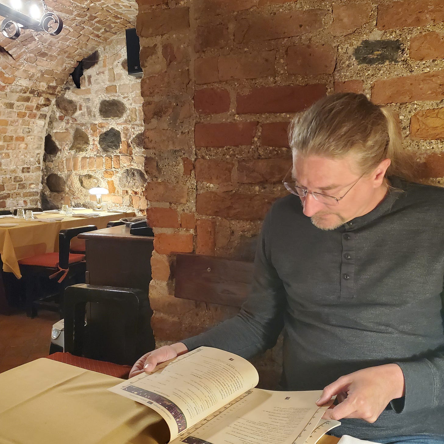 A white man with long blond hair ponders a menu while sitting in a brick alcove.