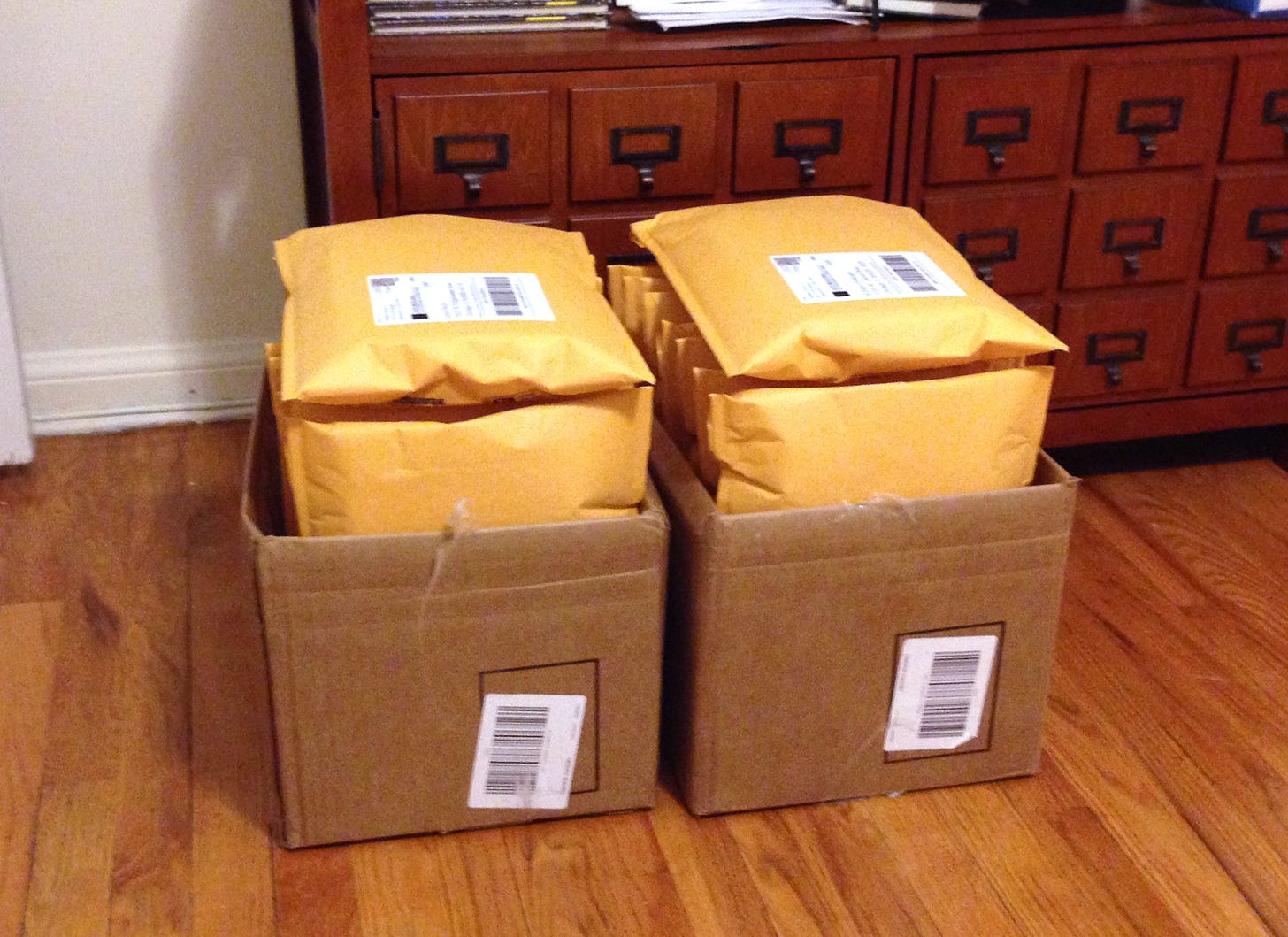 Two cardboard boxes filled with padded yellow book mailers stamped and ready to ship sit on a hardwood before an apothecary-style cabinet.