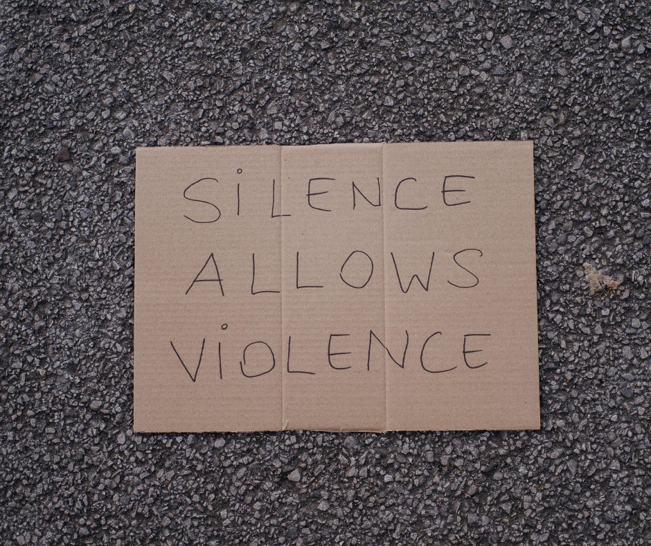 A square piece of brown corrugated cardboard on an asphalt backdrop. On the cardboard, the words "Silence Allows Violence" are handwritten in capital letters using black ink.