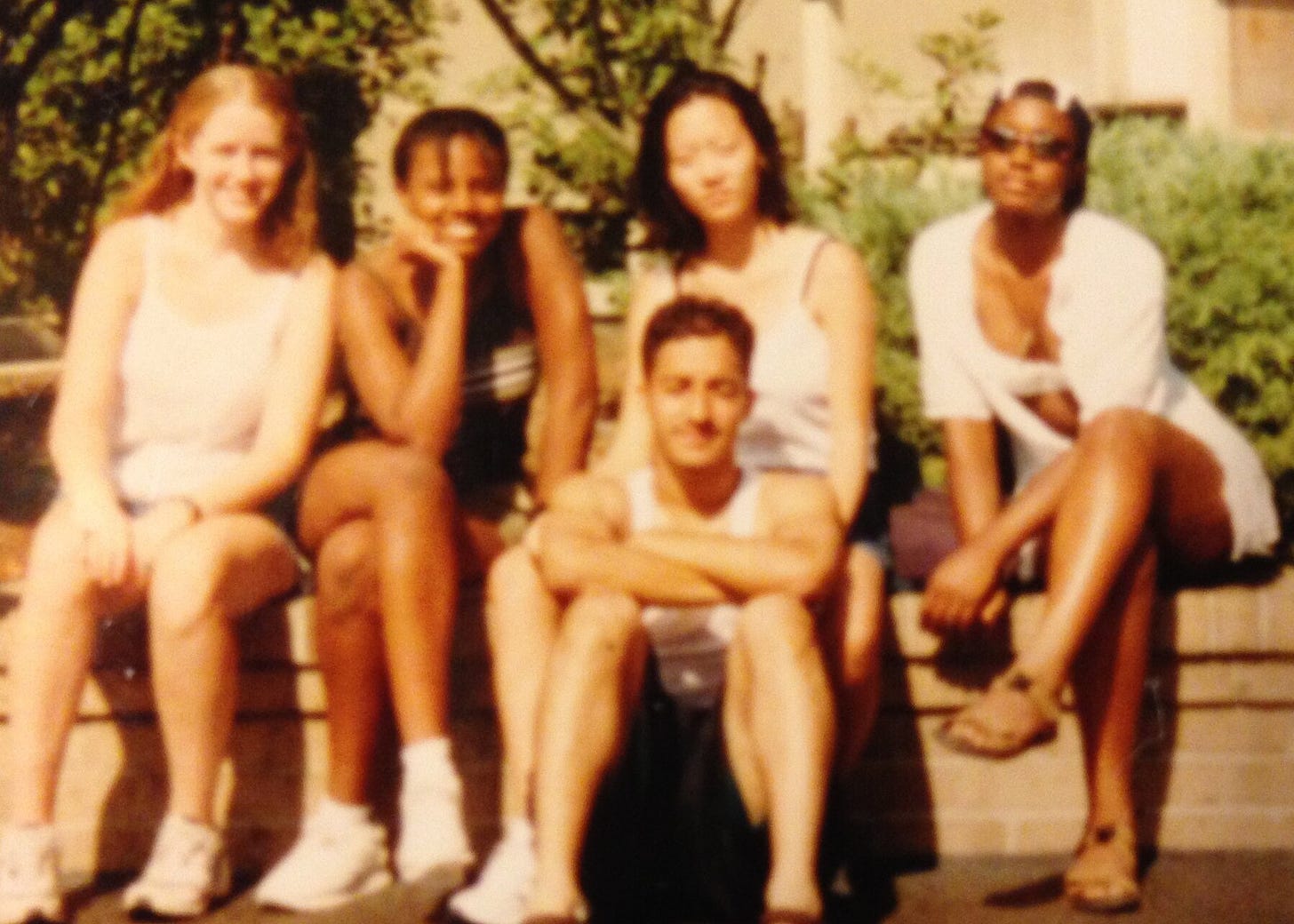 Five teenagers (two Black girls, a white girl, a Korean girl, and a Pakistani boy) sit on a ledge wearing summer clothes